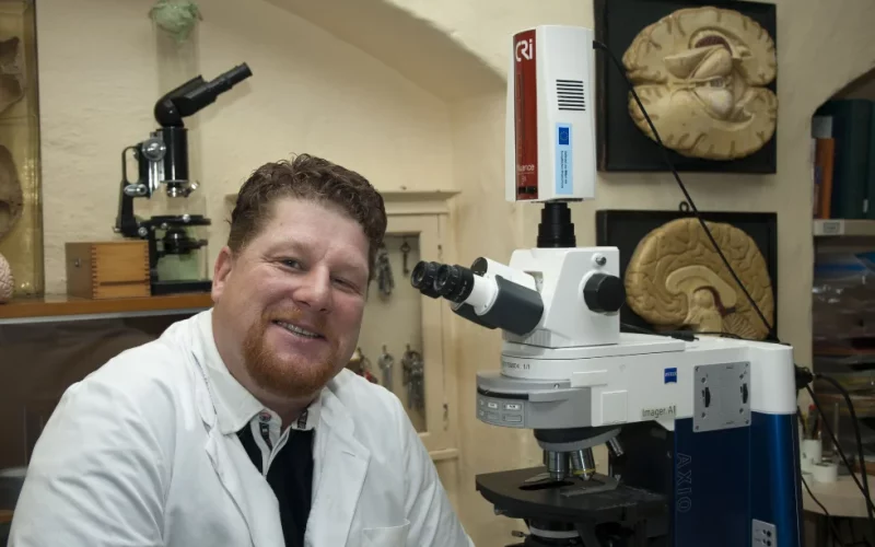 Prof. Dr. Dr. Jens Pahnke in his laboratory in front of a microscope. Illustrations of the human brain are visible on the wall in the background.
